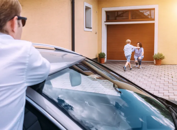 A father watches from his car as his young son runs to hug his mother in front of their home.